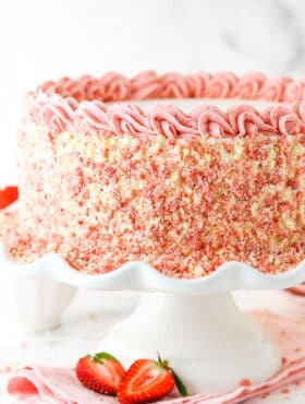 Side view of a full Strawberry Crunchie Ice Cream Cake with pink swirls on a white cake stand