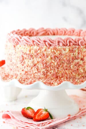 Side view of a full Strawberry Crunchie Ice Cream Cake with pink swirls on a white cake stand