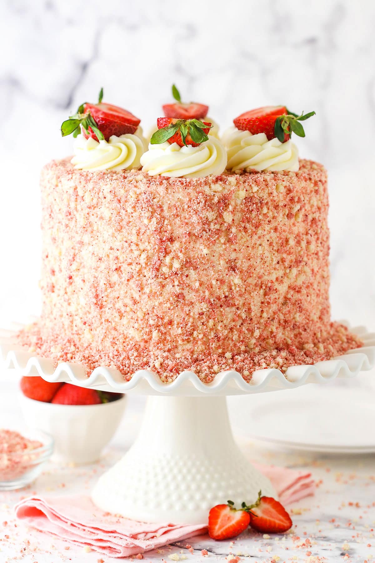 This Strawberry Crunchy Layer Cake is an amazingly delicious cake with layers of vanilla and strawberry cake, strawberry and vanilla frosting and strawberry crunchies on the outside. Strawberry Crunchy Layer Cake with white swirls and cut strawberries on a white cake stand