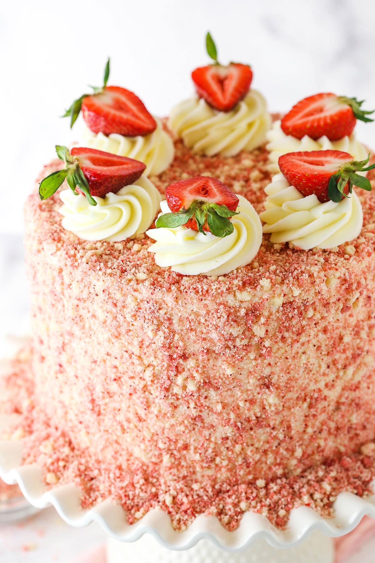 This Strawberry Crunchy Layer Cake is an amazingly delicious cake with layers of vanilla and strawberry cake, strawberry and vanilla frosting and strawberry crunchies on the outside. Strawberry Crunchy Layer Cake with white swirls and cut strawberries on a white cake stand