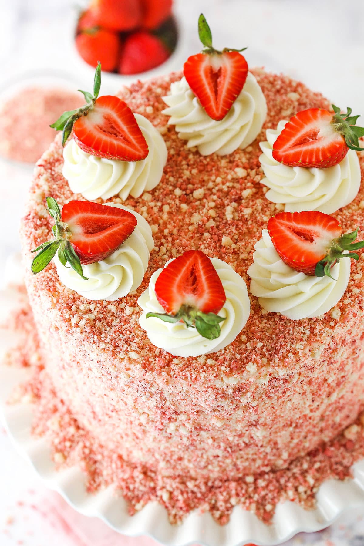 This Strawberry Crunchy Layer Cake is an amazingly delicious cake with layers of vanilla and strawberry cake, strawberry and vanilla frosting and strawberry crunchies on the outside. Strawberry Crunchy Layer Cake with white swirls and cut strawberries on a white cake stand