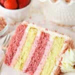 A slice of Strawberry Crunchy Layer Cake on a white plate with a fork and strawberries in the background