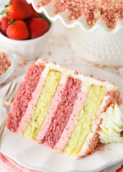 A slice of Strawberry Crunchy Layer Cake on a white plate with a fork and strawberries in the background