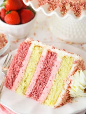 A slice of Strawberry Crunchy Layer Cake on a white plate with a fork and strawberries in the background