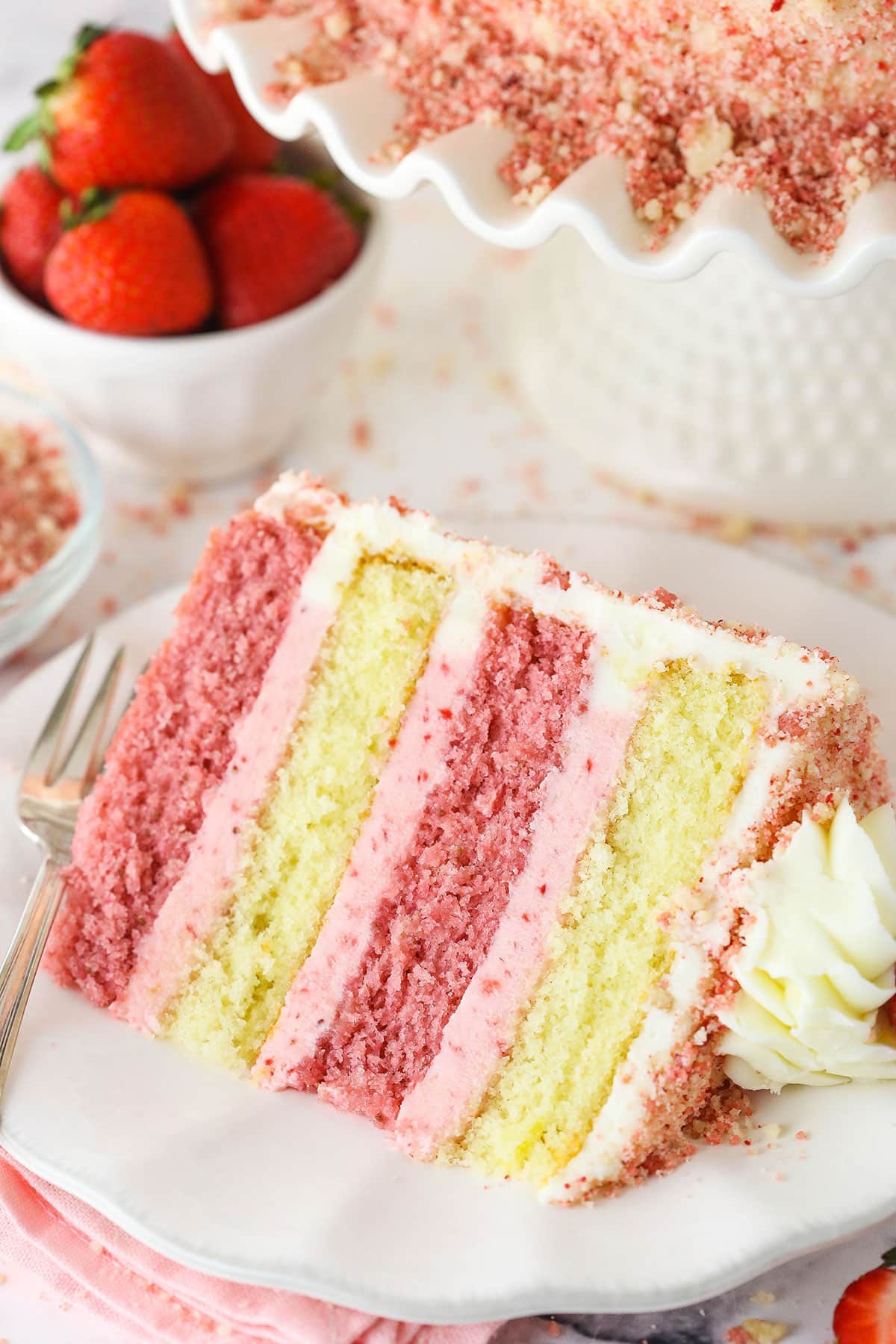 A slice of Strawberry Crunchy Layer Cake on a white plate with a fork and strawberries in the background