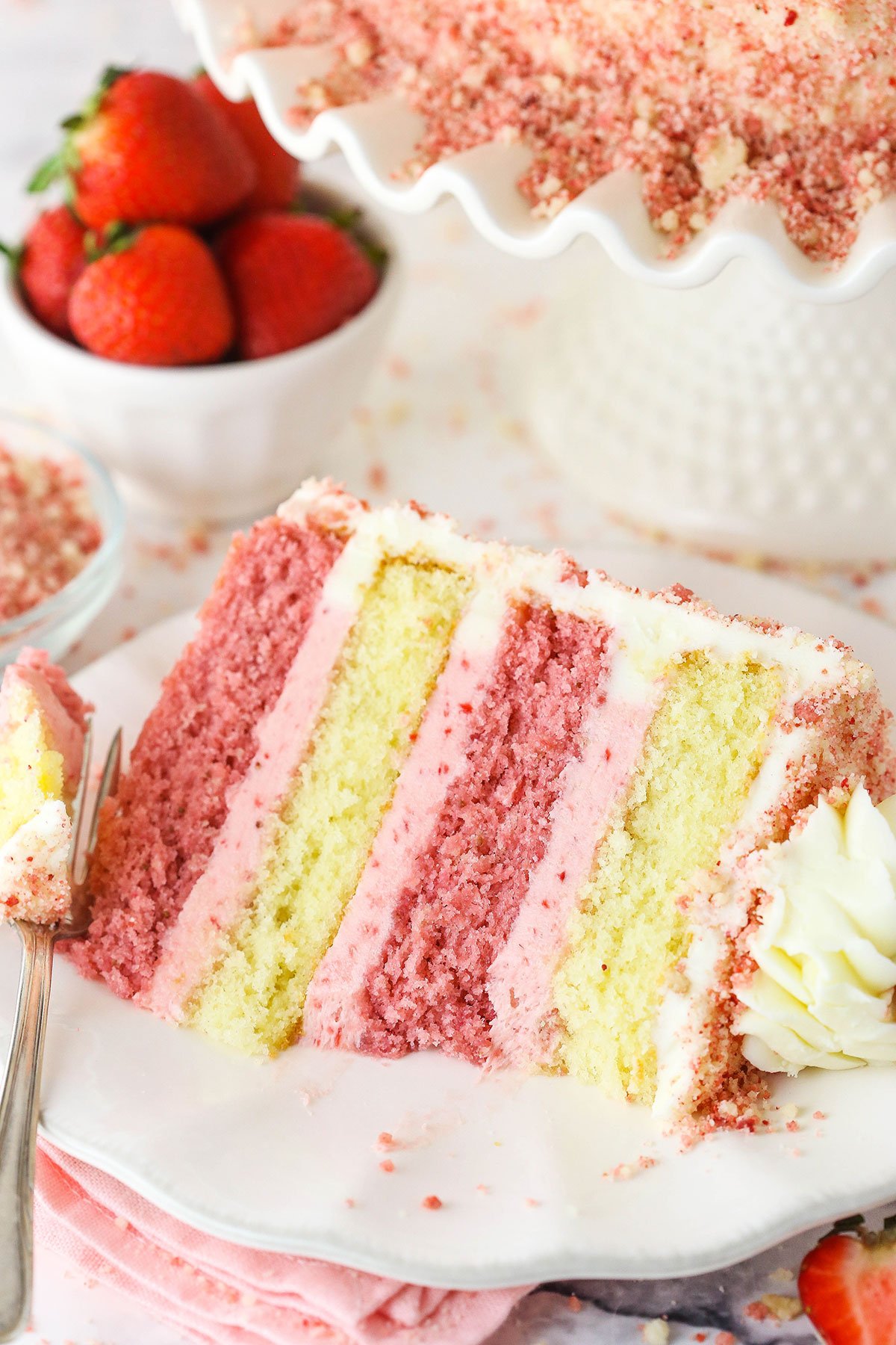 This Strawberry Crunchy Layer Cake is an amazingly delicious cake with layers of vanilla and strawberry cake, strawberry and vanilla frosting and strawberry crunchies on the outside. A slice of Strawberry Crunchy Layer Cake with a bite taken out on a white plate with a fork and strawberries in the background