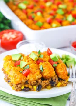 A serving of Taco Tater Tot Casserole topped with chopped avocado and tomato on a white plate with a fork