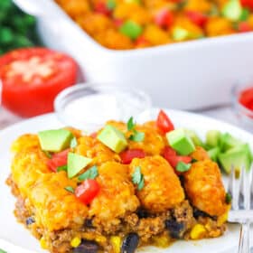 A serving of Taco Tater Tot Casserole topped with chopped avocado and tomato on a white plate with a fork
