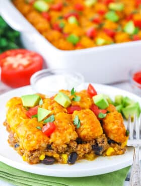 A serving of Taco Tater Tot Casserole topped with chopped avocado and tomato on a white plate with a fork