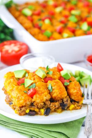 A serving of Taco Tater Tot Casserole topped with chopped avocado and tomato on a white plate with a fork