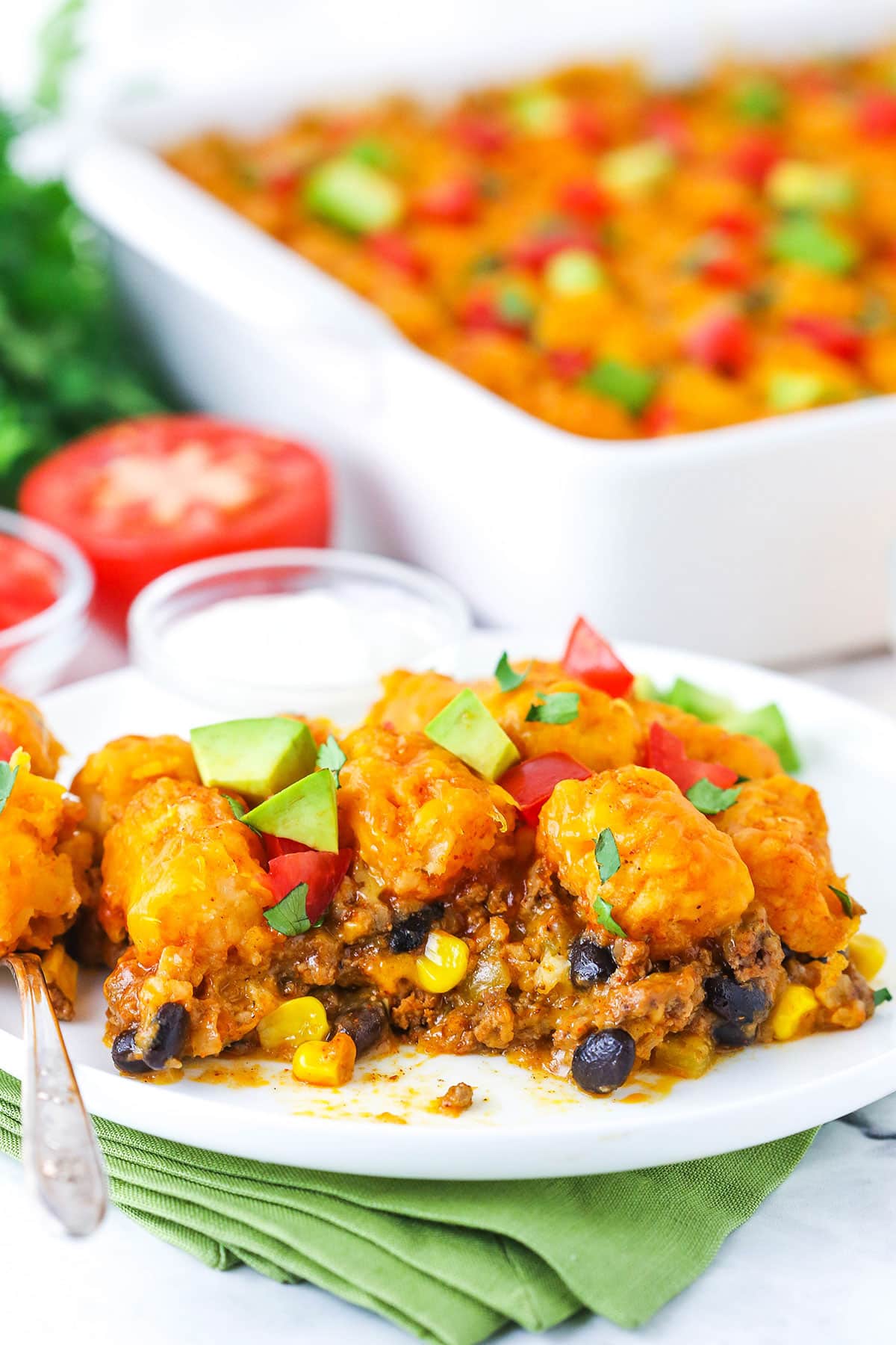 A serving of Taco Tater Tot Casserole topped with chopped avocado and tomato on a white plate with a fork