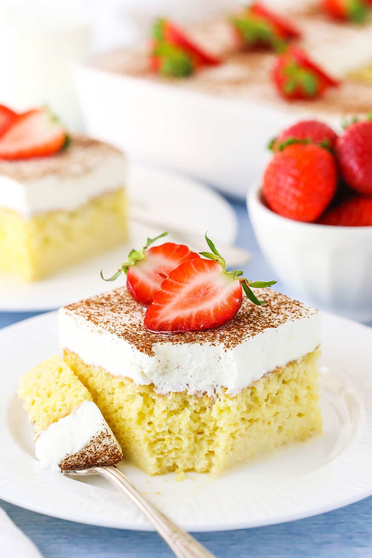 A square serving of Tres Leches Cake with a bite taken out with cut strawberries on top on a white plate with a fork