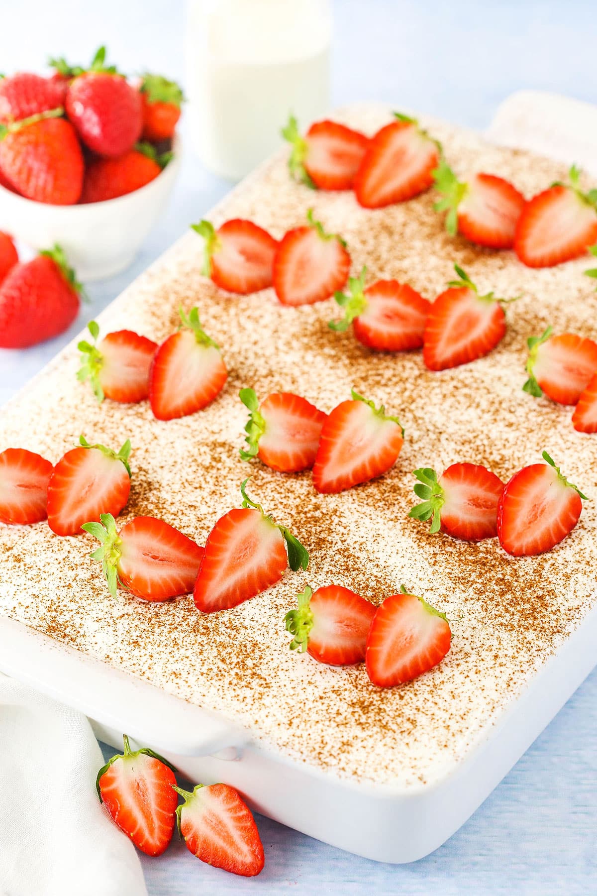 A full Tres Leches Cake topped with cut strawberries in a white platter with whole strawberries in the background