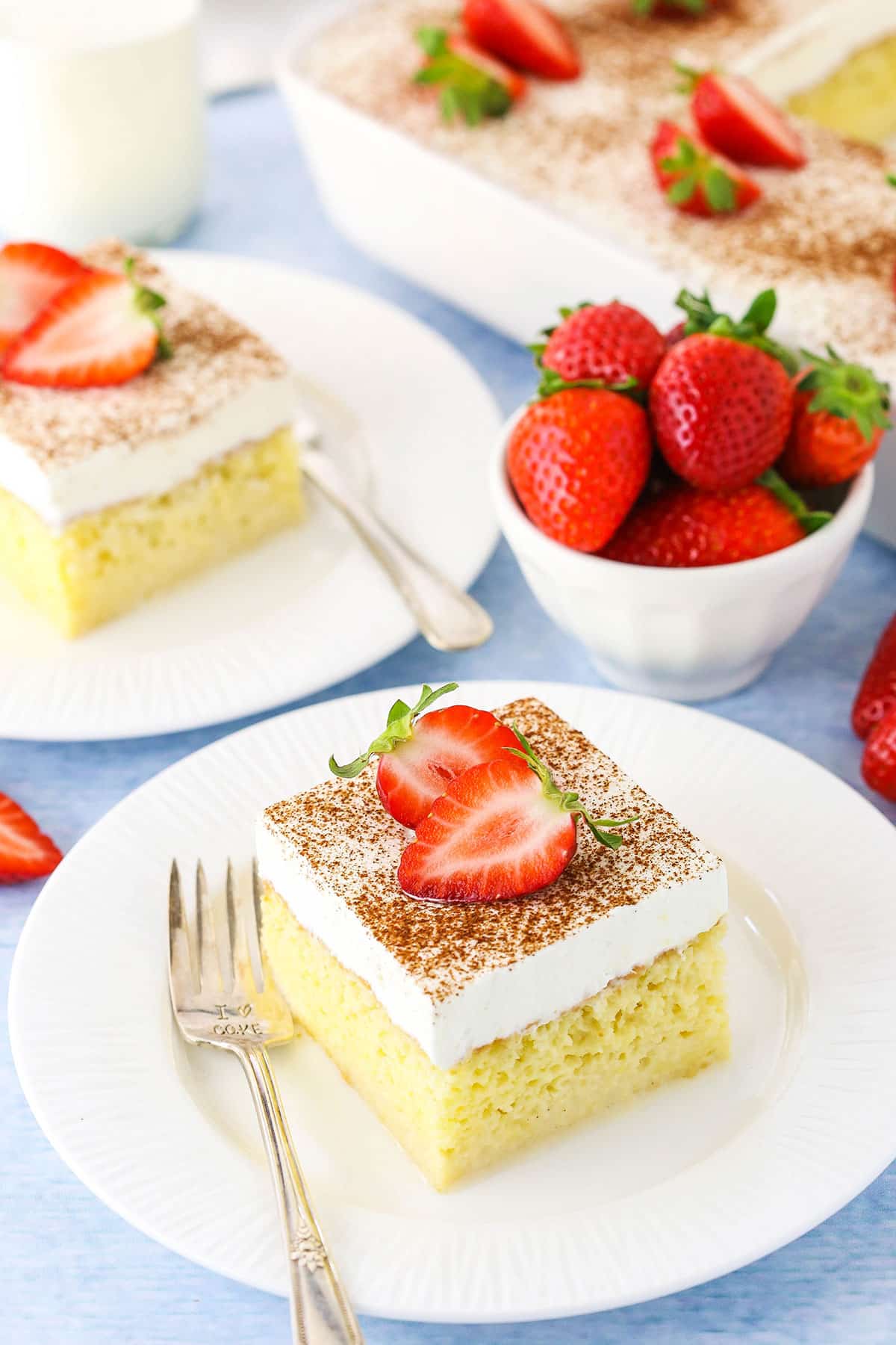 A square serving of Tres Leches Cake with cut strawberries on top on a white plate with a fork