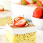 A square serving of Tres Leches Cake with cut strawberries on top on a white plate with a fork