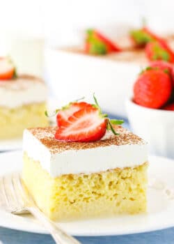 A square serving of Tres Leches Cake with cut strawberries on top on a white plate with a fork