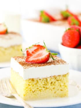 A square serving of Tres Leches Cake with cut strawberries on top on a white plate with a fork