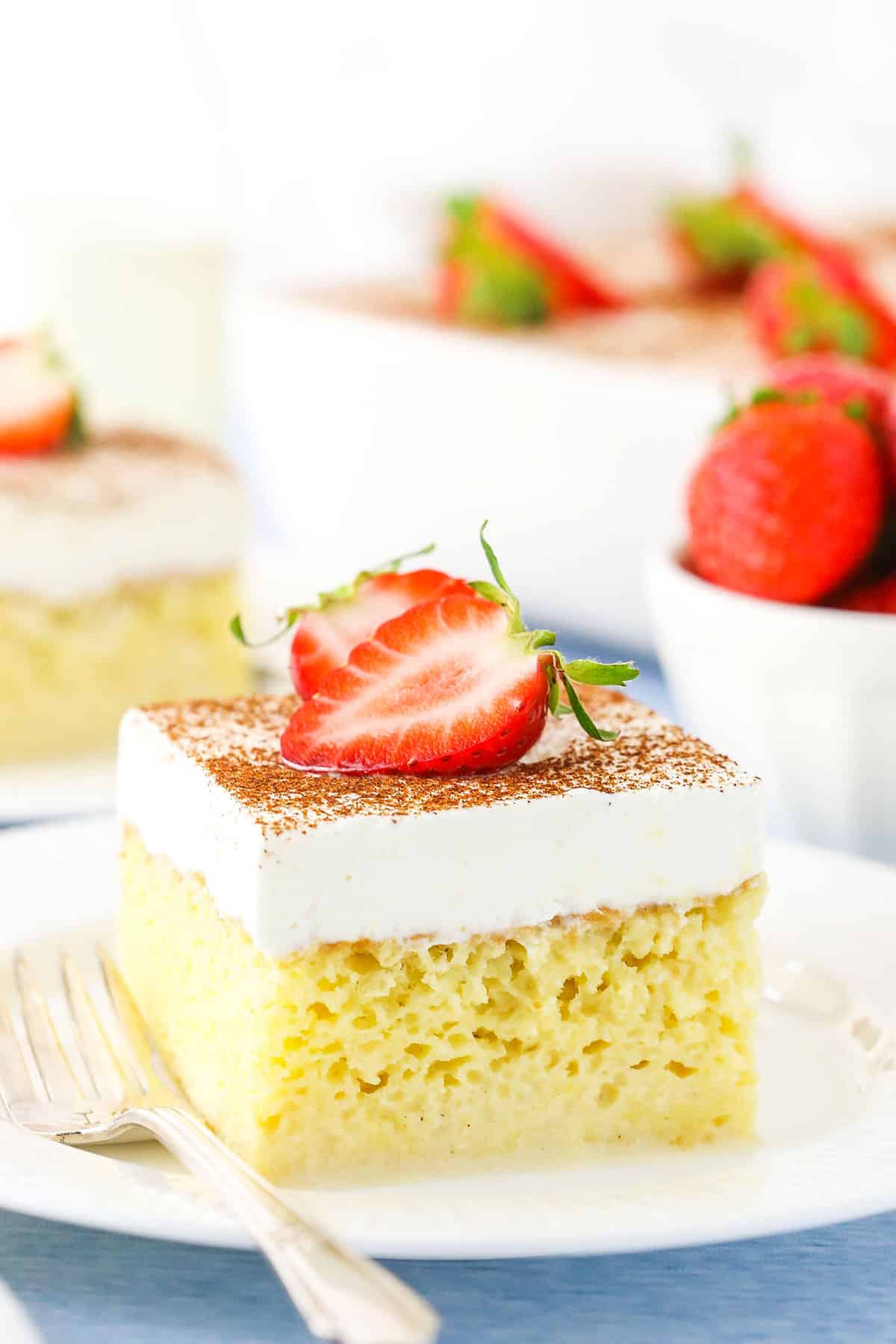 A square serving of Tres Leches Cake with cut strawberries on top on a white plate with a fork