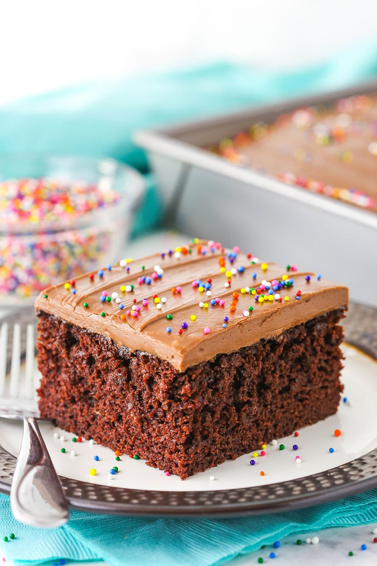 A single square serving of Wacky Cake with chocolate frosting and sprinkles on a white plate with a fork