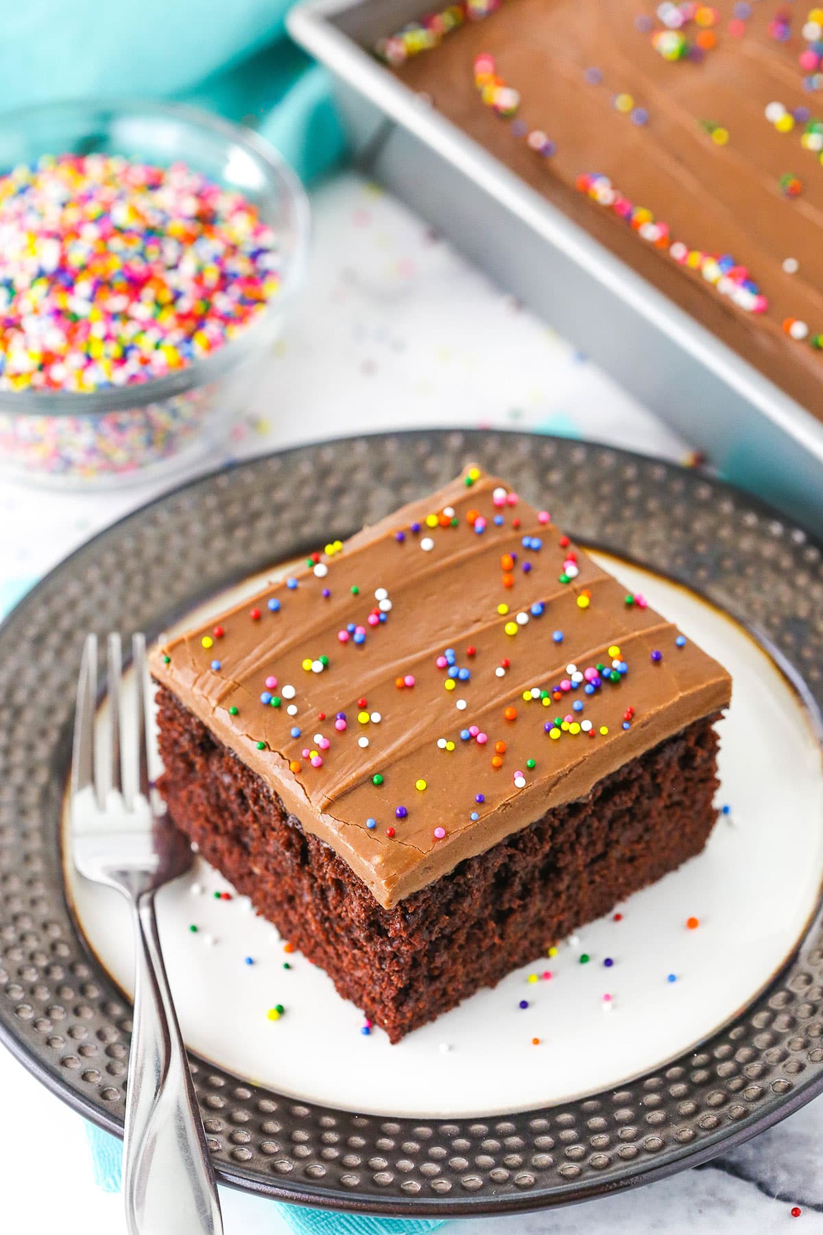 Overhead view of a single square serving of Wacky Cake with chocolate frosting and sprinkles on a white plate with a fork