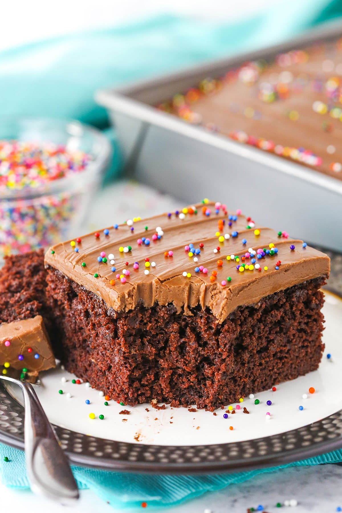 A single square serving of Wacky Cake with chocolate frosting and sprinkles with a bite removed on a white plate with a fork