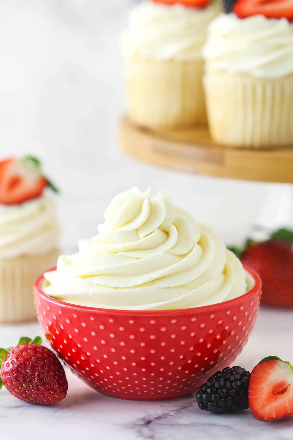 Whipped cream frosting in a decorative bowl near frosted cupcake surrounded by fresh berries.