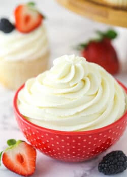 Whipped cream frosting in a decorative bowl near frosted cupcake surrounded by fresh berries.