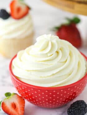 Whipped cream frosting in a decorative bowl near frosted cupcake surrounded by fresh berries.