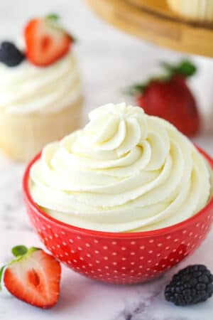 Whipped cream frosting in a decorative bowl near frosted cupcake surrounded by fresh berries.