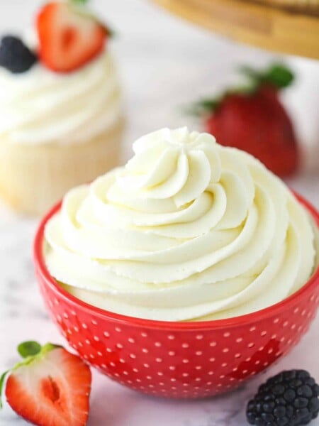 Whipped cream frosting in a decorative bowl near frosted cupcake surrounded by fresh berries.