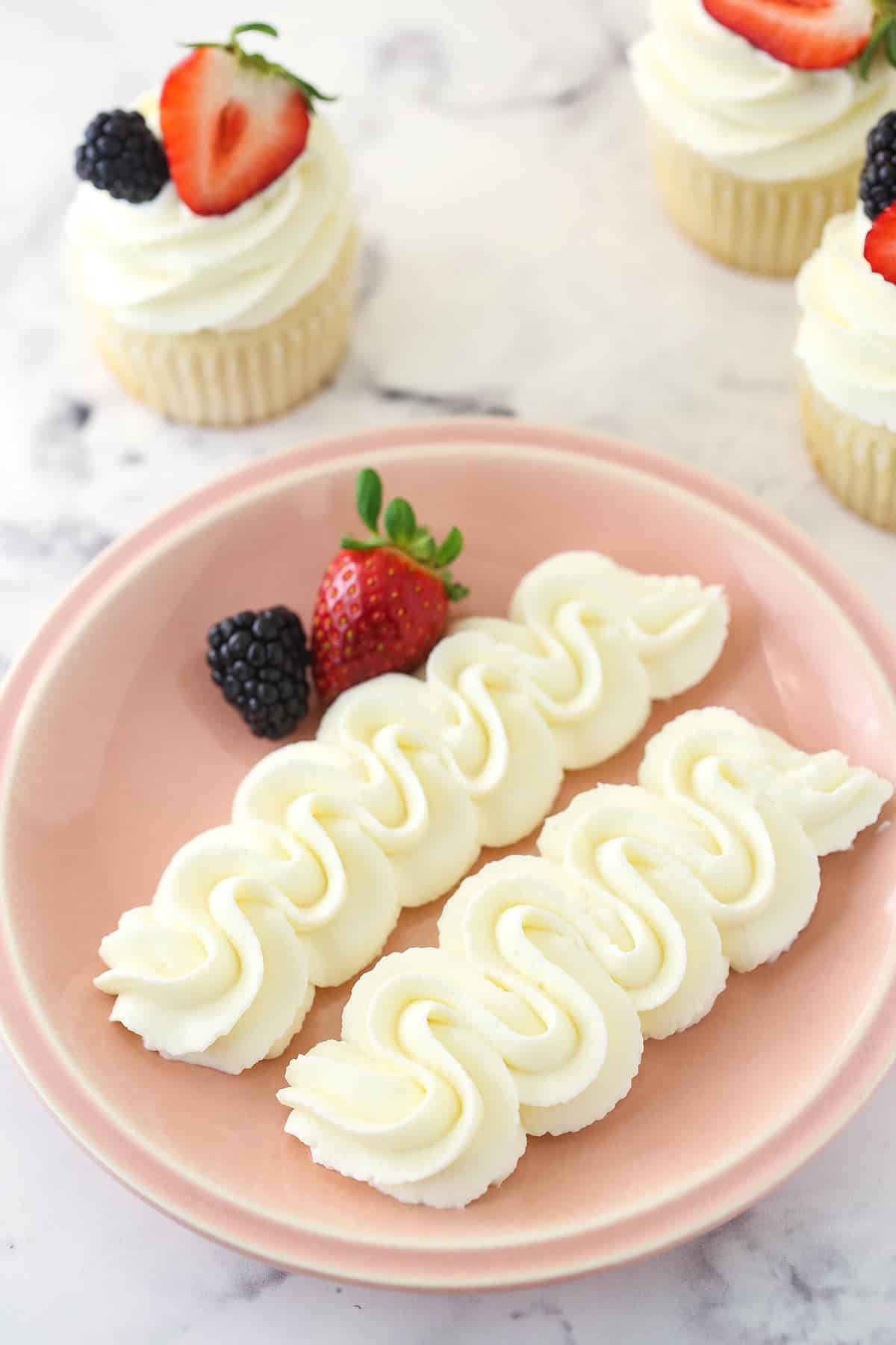 Whipped cream cheese frosting piped onto a plate with fresh berries surrounded by frosted cupcakes.