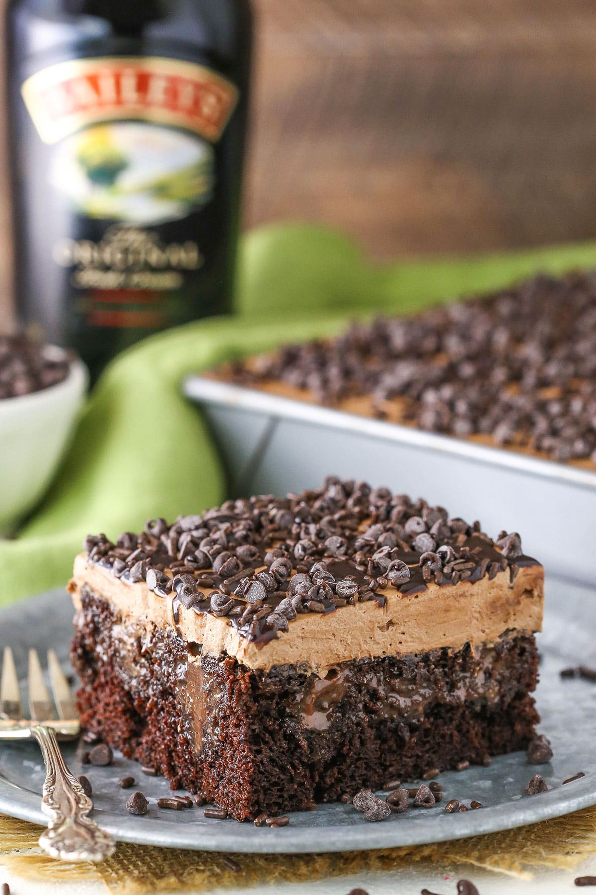 A square slice of Baileys Chocolate Poke Cake topped with chocolate chips on a silver plate with a fork and a bottle of Baileys in the background
