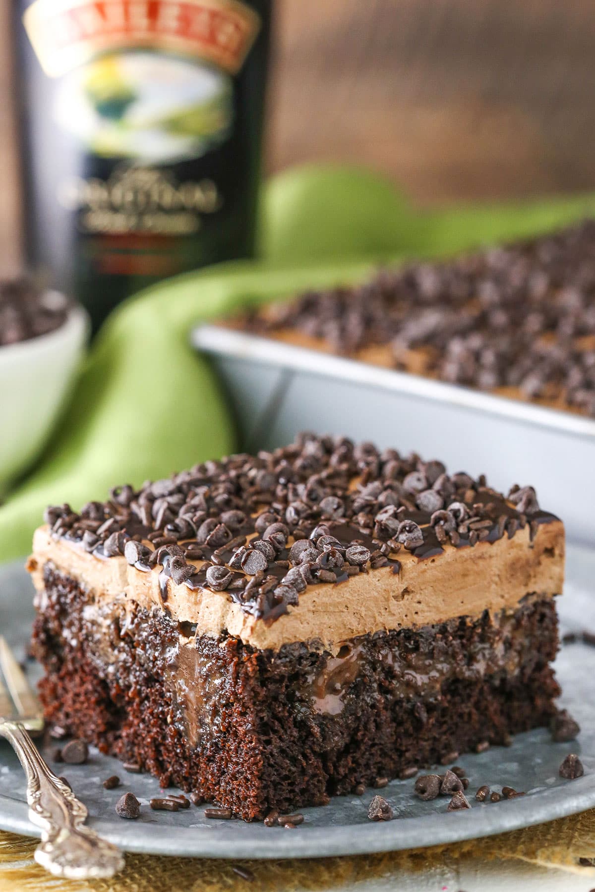 A square slice of Baileys Chocolate Poke Cake topped with chocolate chips on a silver plate with a fork and a bottle of Baileys in the background