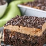 A square slice of Baileys Chocolate Poke Cake topped with chocolate chips on a silver plate with a fork and a bottle of Baileys in the background