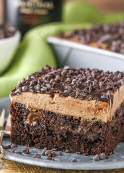 A square slice of Baileys Chocolate Poke Cake topped with chocolate chips on a silver plate with a fork and a bottle of Baileys in the background