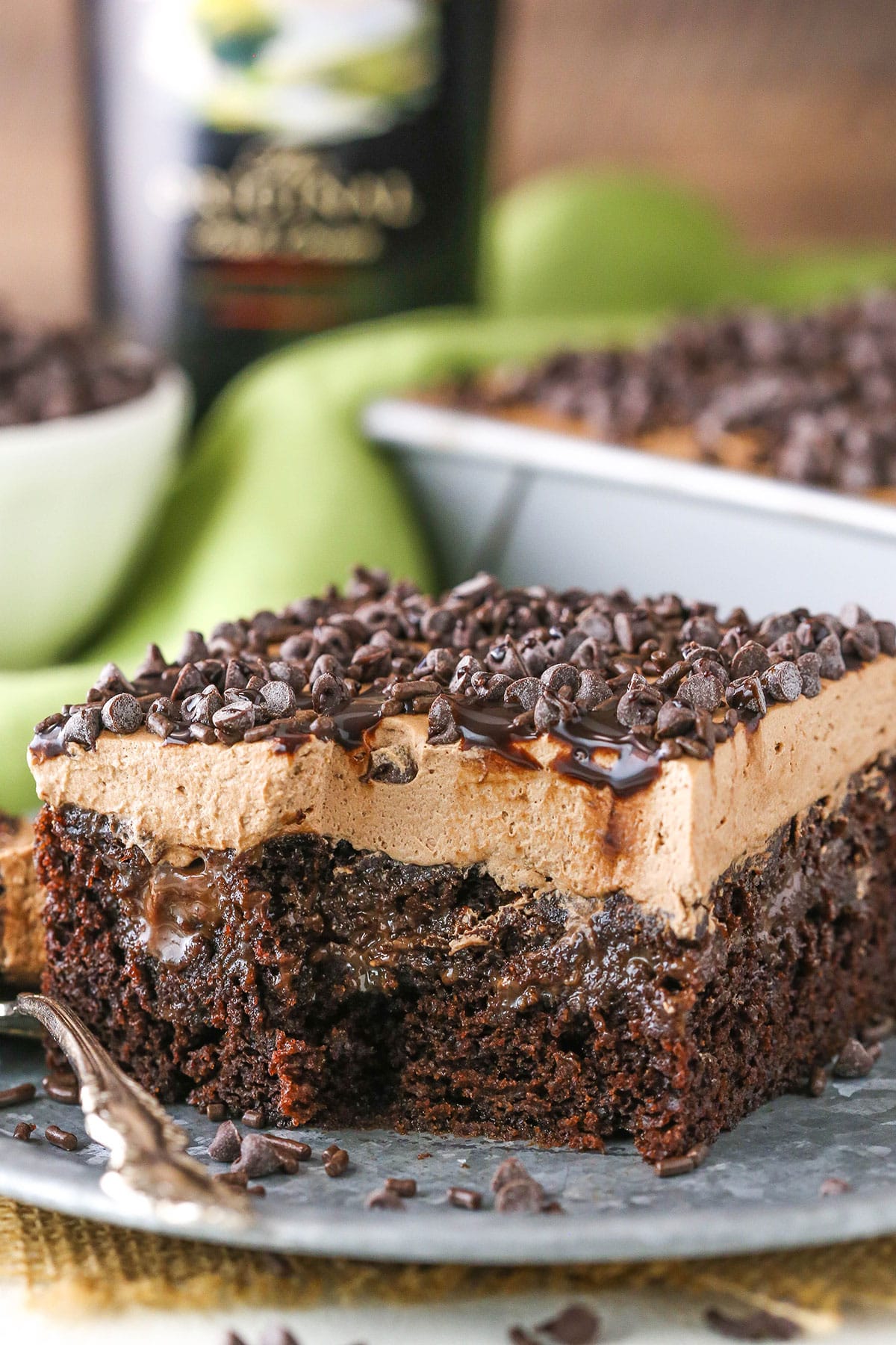 A square slice of Baileys Chocolate Poke Cake topped with chocolate chips with a bite taken out on a silver plate with a fork and a bottle of Baileys in the background