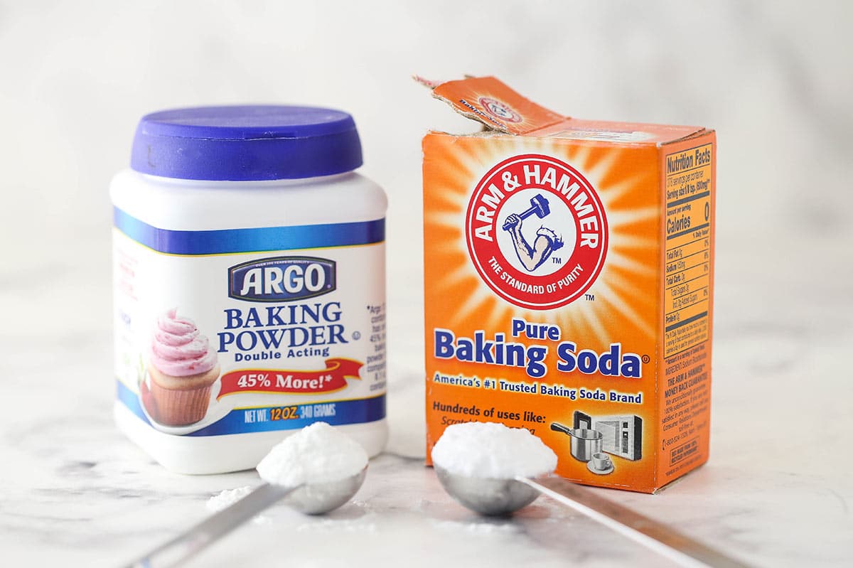 a container of baking soda and baking powder sitting on marble background with a scoop of each in front