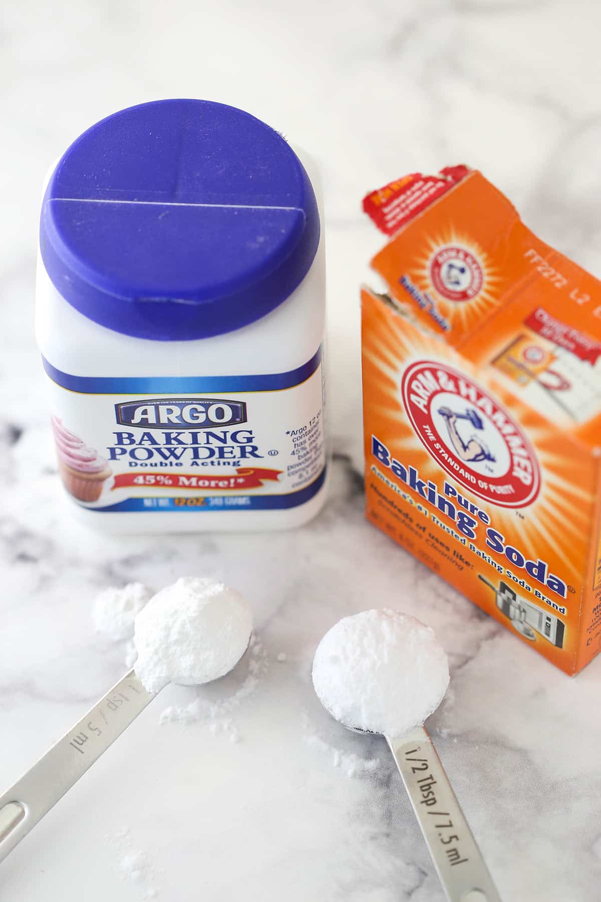 overhead image of a container of baking soda and baking powder sitting on marble background with a scoop of each in front