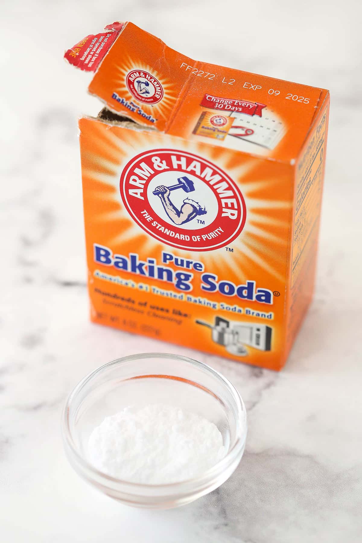 a box of baking soda with a little bit in a glass bowl in front on marble background