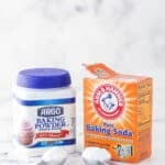 a container of baking soda and baking powder sitting on marble background with a scoop of each in front