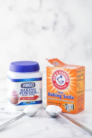 a container of baking soda and baking powder sitting on marble background with a scoop of each in front