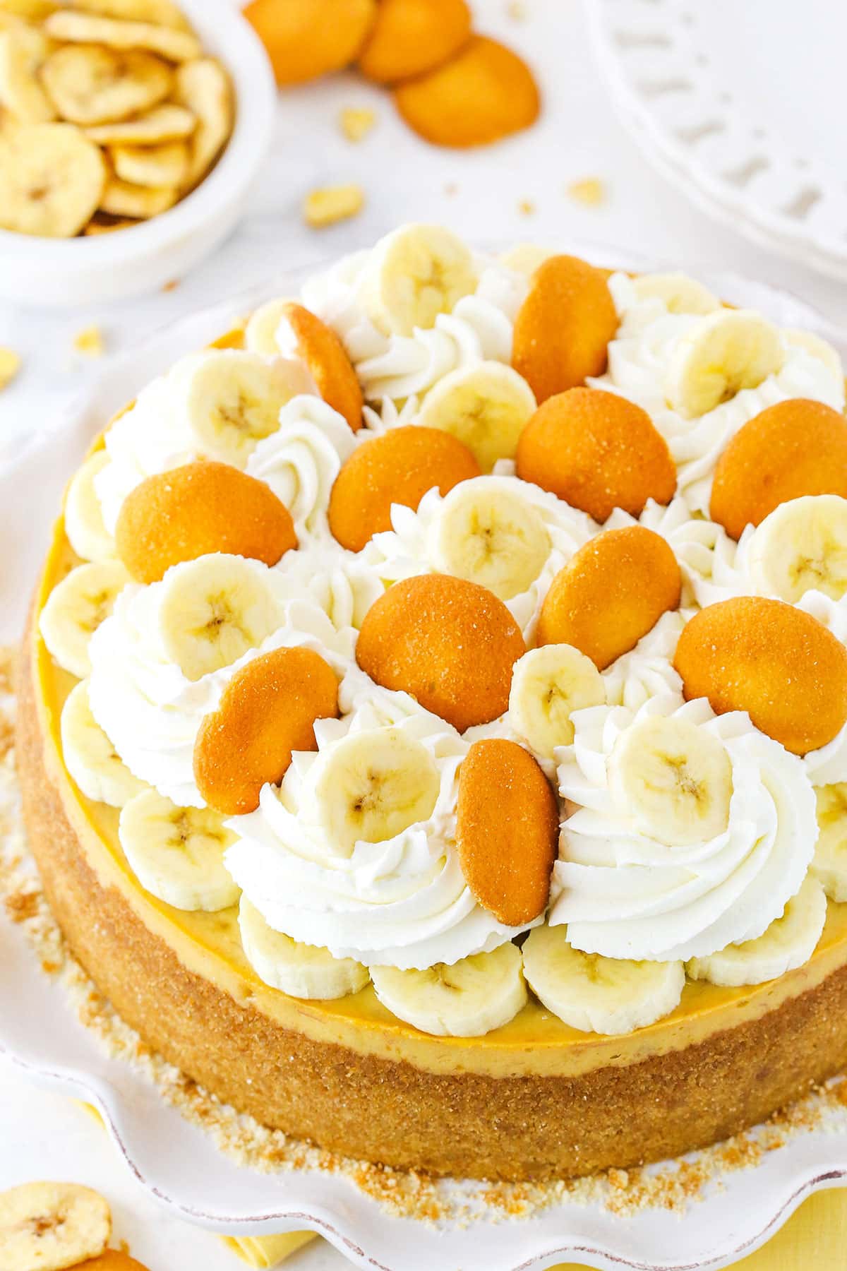 This Banana Pudding Cheesecake is made with a vanilla wafer crust, loads of mashed and sliced bananas, and banana pudding mix! It’s got the best banana pudding flavor you could have in a cheesecake and it’s completely addicting! Overhead view of a full Banana Pudding Cheesecake topped with whipped cream, vanilla wafers and sliced banana on a white platter