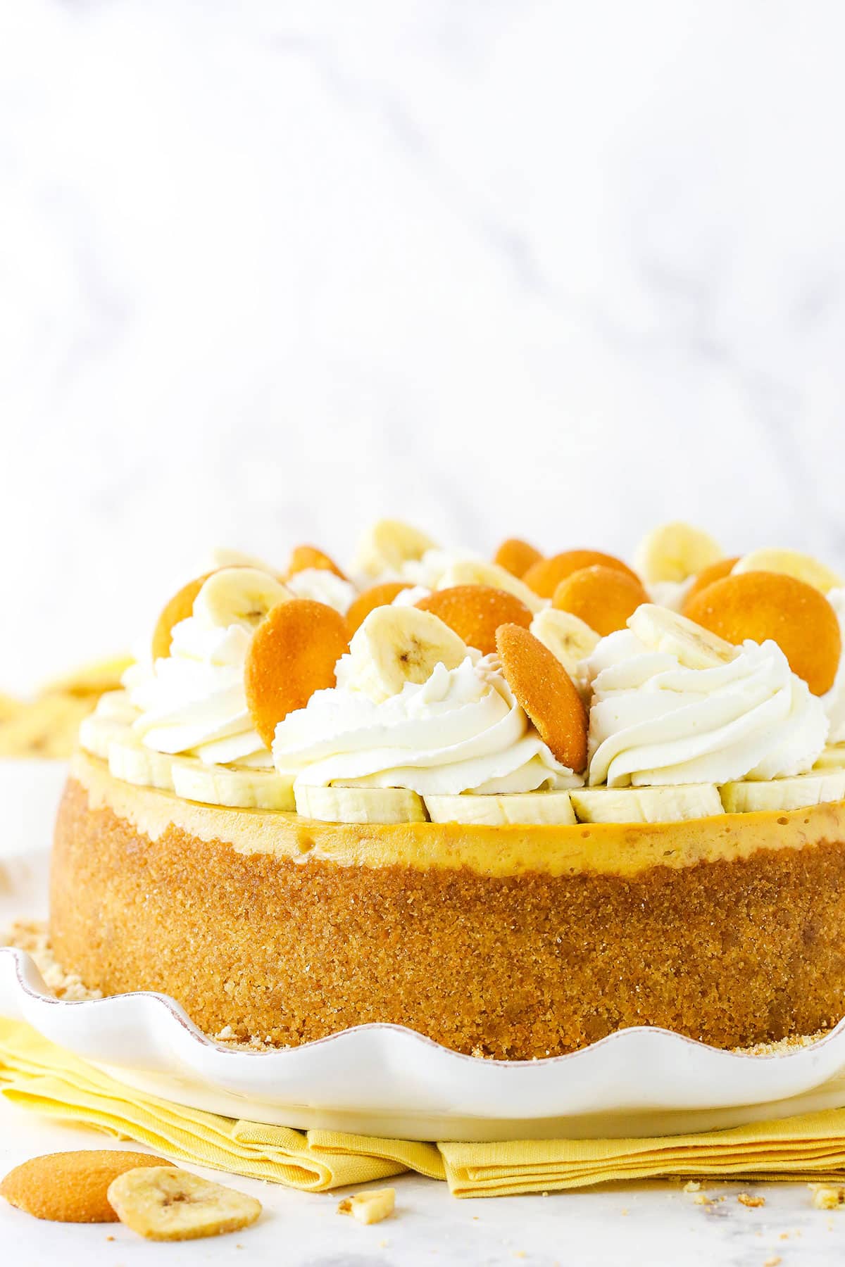 Side view of a full Banana Pudding Cheesecake topped with whipped cream, vanilla wafers and sliced banana on a white platter