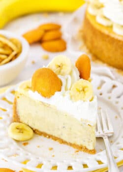 A slice of Banana Pudding Cheesecake topped with whipped cream, vanilla wafers and sliced banana on a white plate next to a fork