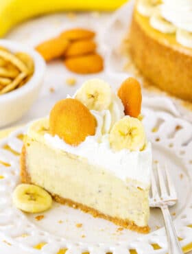 A slice of Banana Pudding Cheesecake topped with whipped cream, vanilla wafers and sliced banana on a white plate next to a fork