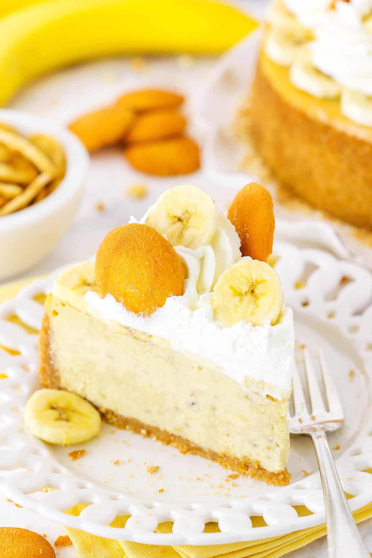 A slice of Banana Pudding Cheesecake topped with whipped cream, vanilla wafers and sliced banana on a white plate next to a fork