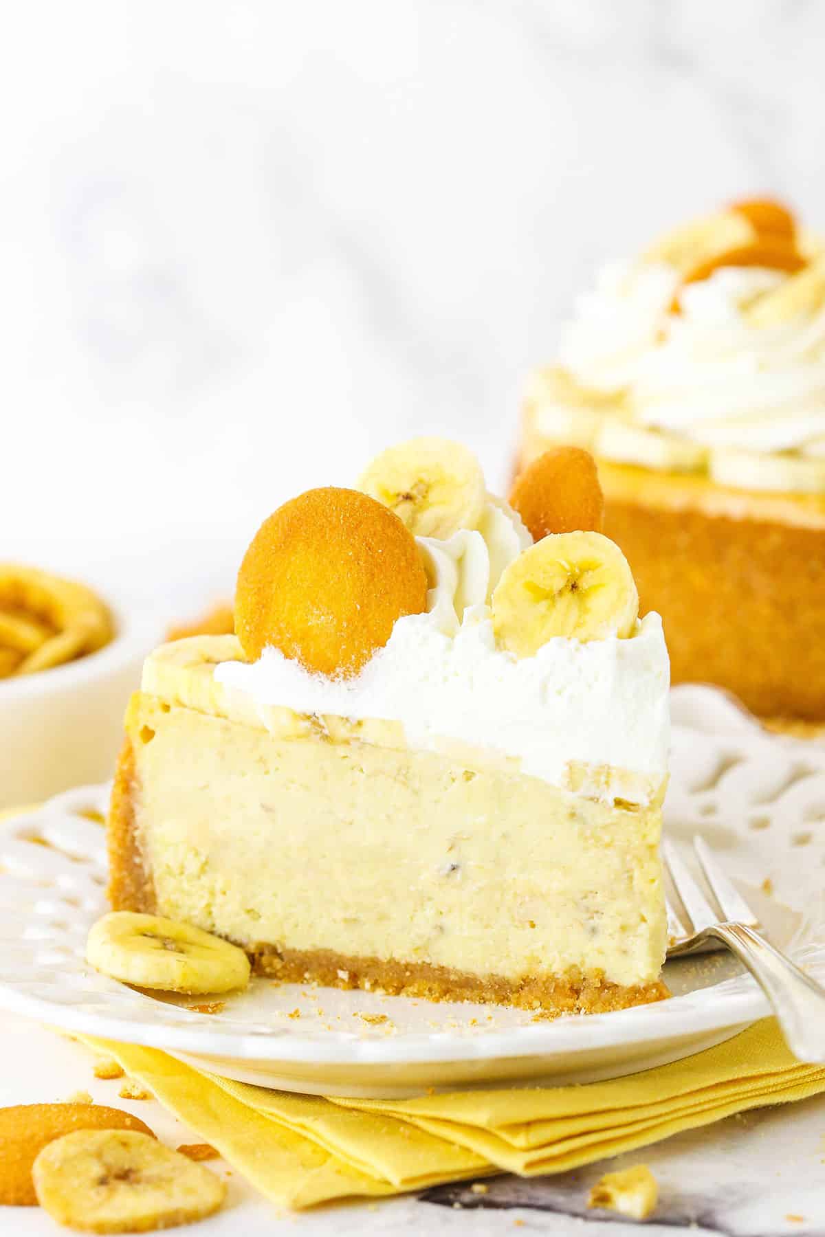 A slice of Banana Pudding Cheesecake topped with whipped cream, vanilla wafers and sliced banana on a white plate next to a fork