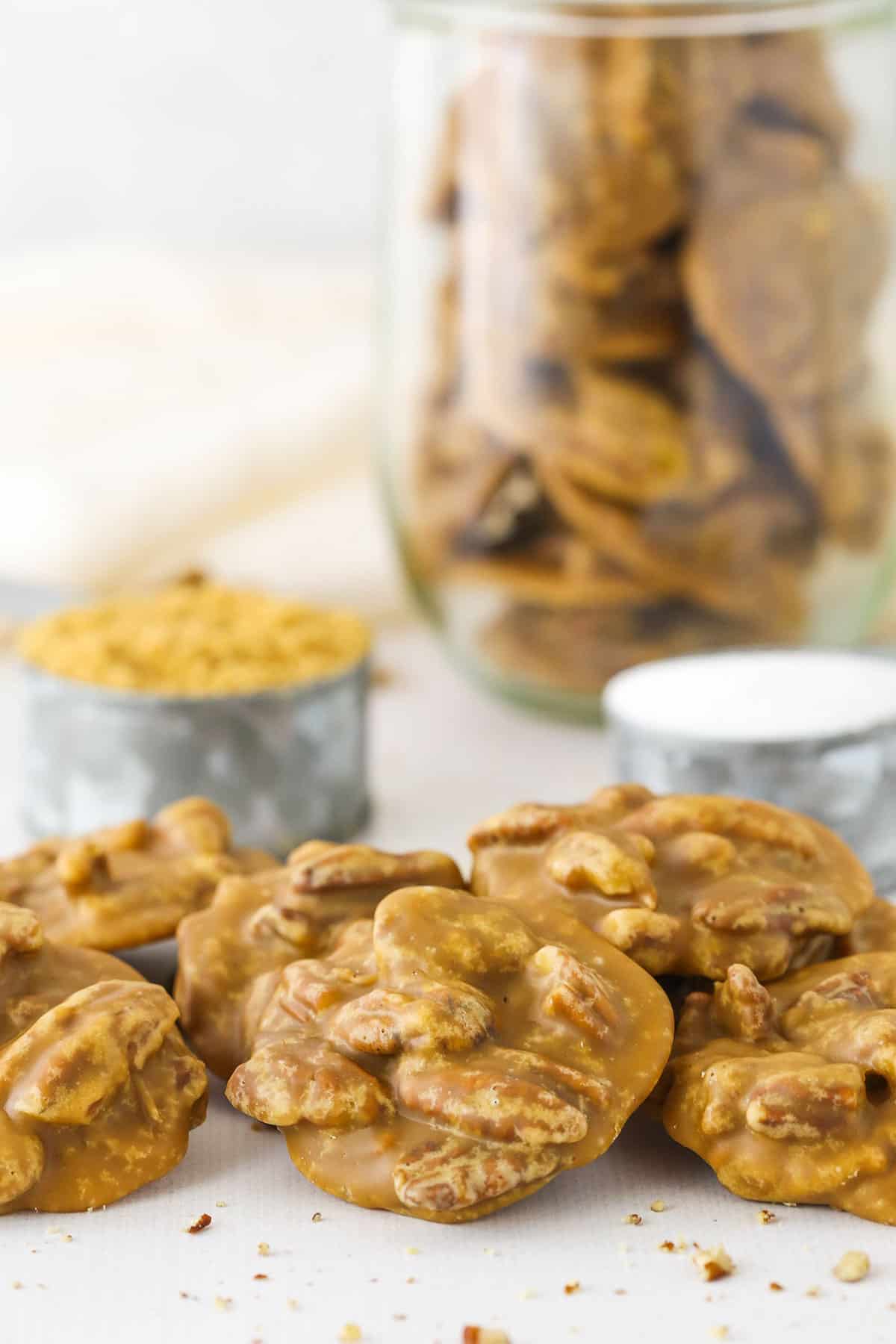 These are seriously the BEST Southern Praline Pecans! With the amazing flavor of brown sugar and butter, these are pure heaven! A little bit candy and a little bit cookie, they melt right in your mouth! Six Southern Pecan Pralines on a white table with brown sugar and a jar of more Southern Pecan Pralines in the background