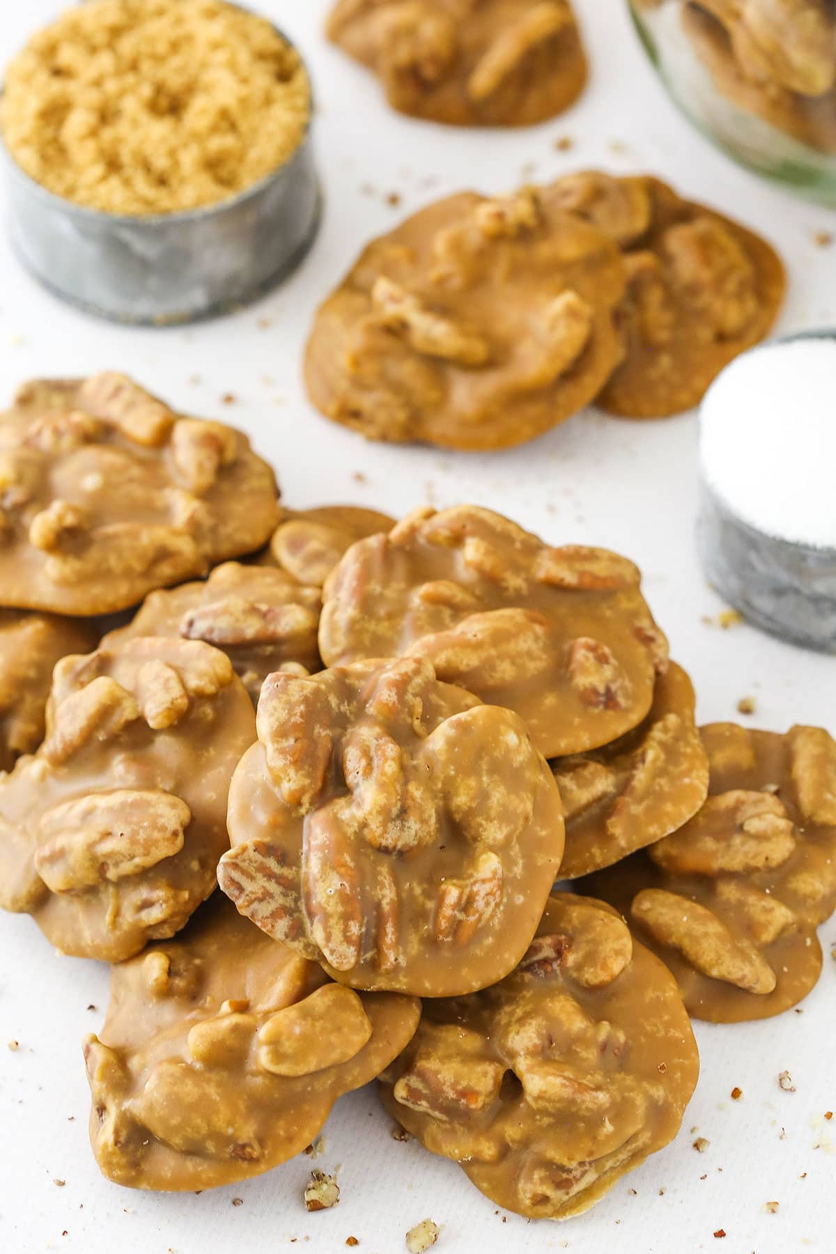 Southern Pecan Pralines layered on a white table with a cup of brown sugar and a cup of sugar in the background