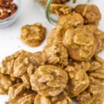 Southern Pecan Pralines poured out of a glass jar on it's side onto a white table with a bowl of pecans in the background